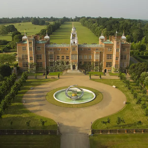 Aerial view of a grand, castle-like mansion with multiple towers, nestled in Hertfordshire's lush greenery and featuring a circular driveway with a central fountain, evoking the charm of historical sites. seo hertfordshire